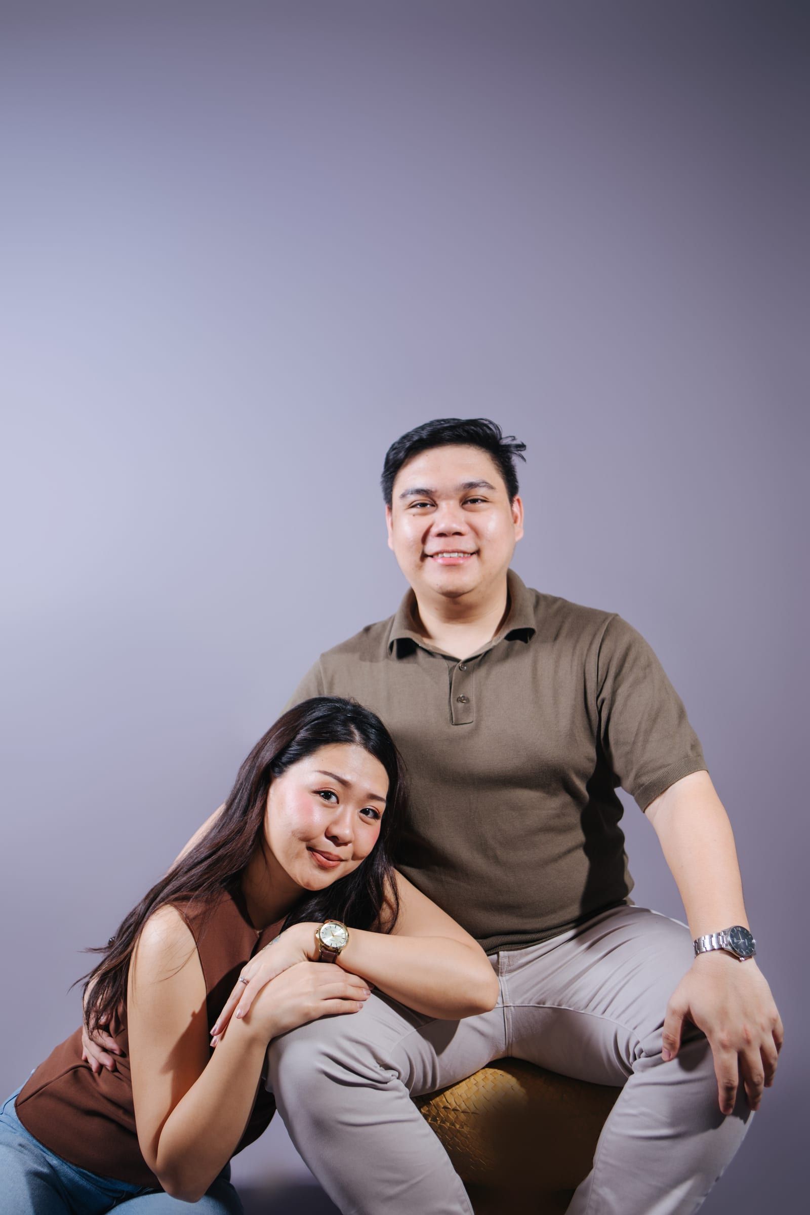 Anna leans her head on Joboy's knee and smiles at the camera while Joboy sits behind her on a rattan stool, against a lavender studio backdrop.