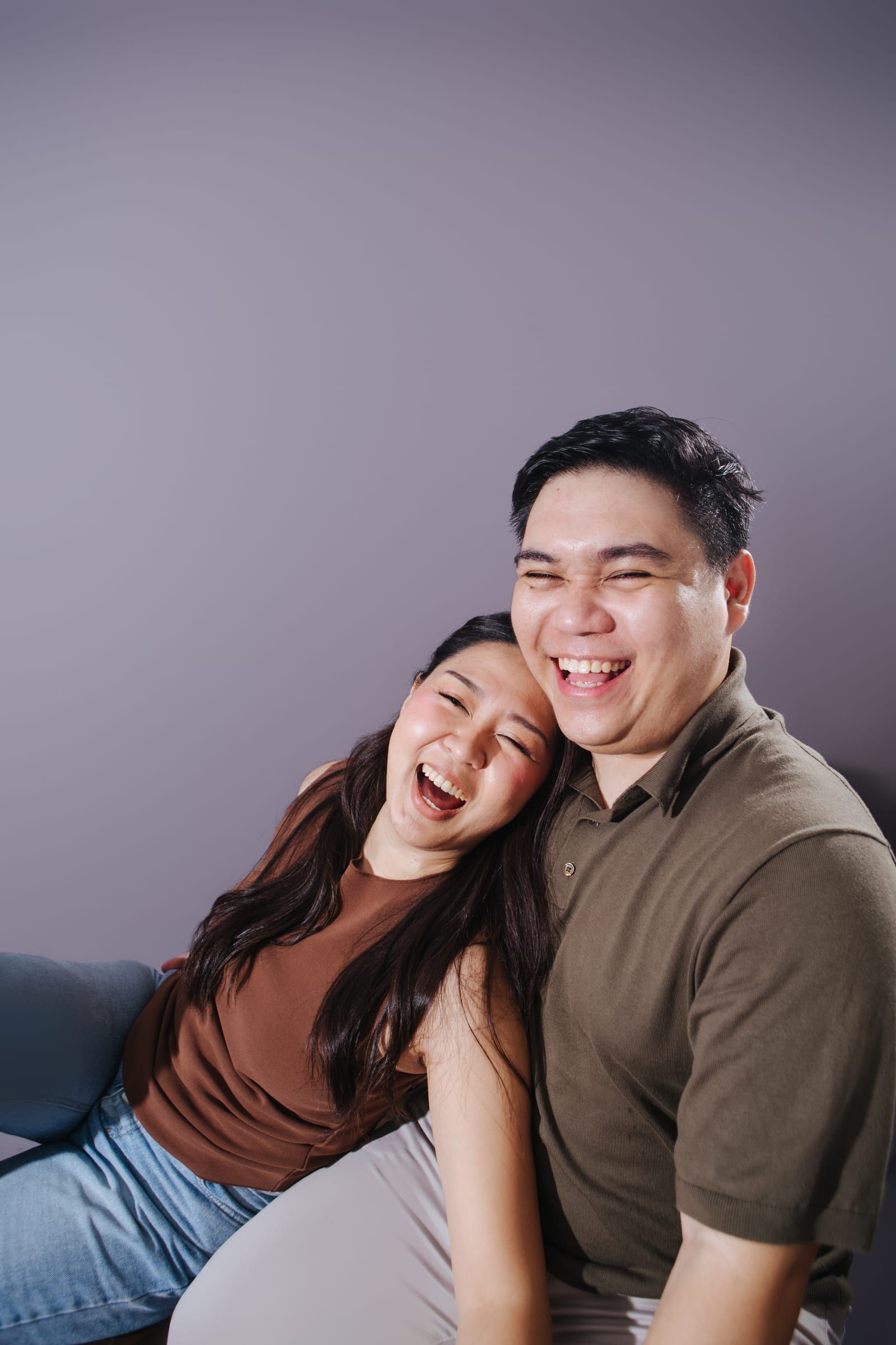 Anna and Joboy laugh with their eyes shut, her head tipped against his shoulder, in front of a lavender backdrop.