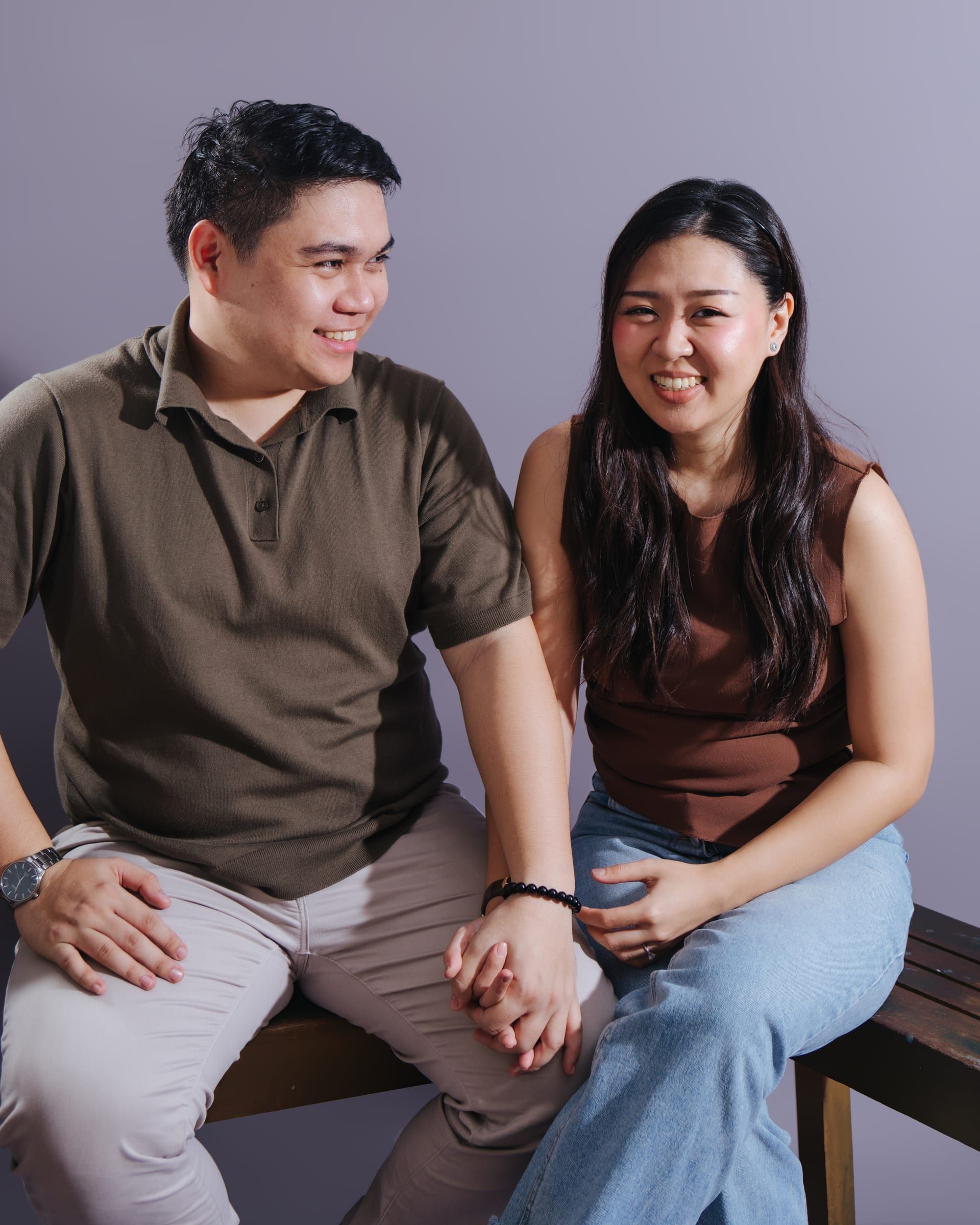 Now: Joboy, in an olive polo and light chinos, and Anna, in a brown sleeveless top and jeans, sit on a wooden bench holding hands against a lavender backdrop; she smiles at the camera and he smiles at her.